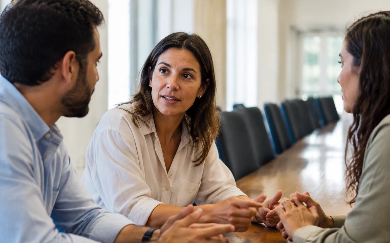 Três participantes conversam ao redor de uma mesa comprida numa sala clara de janelas altas